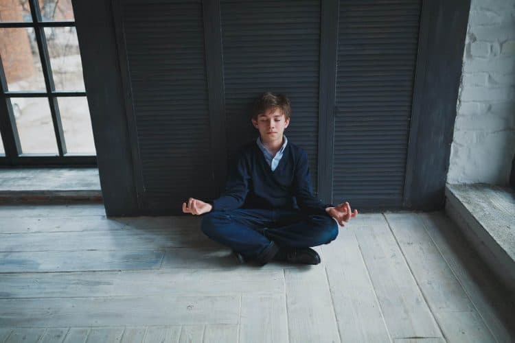 Calm teenager boy sitting on the floor near the dark blue walls in the room in Lotus position