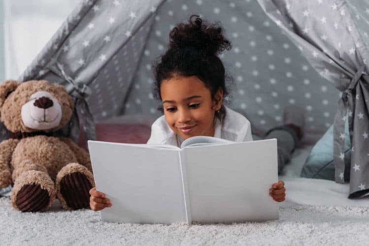 adorable african american kid reading book and lying in tent at home
