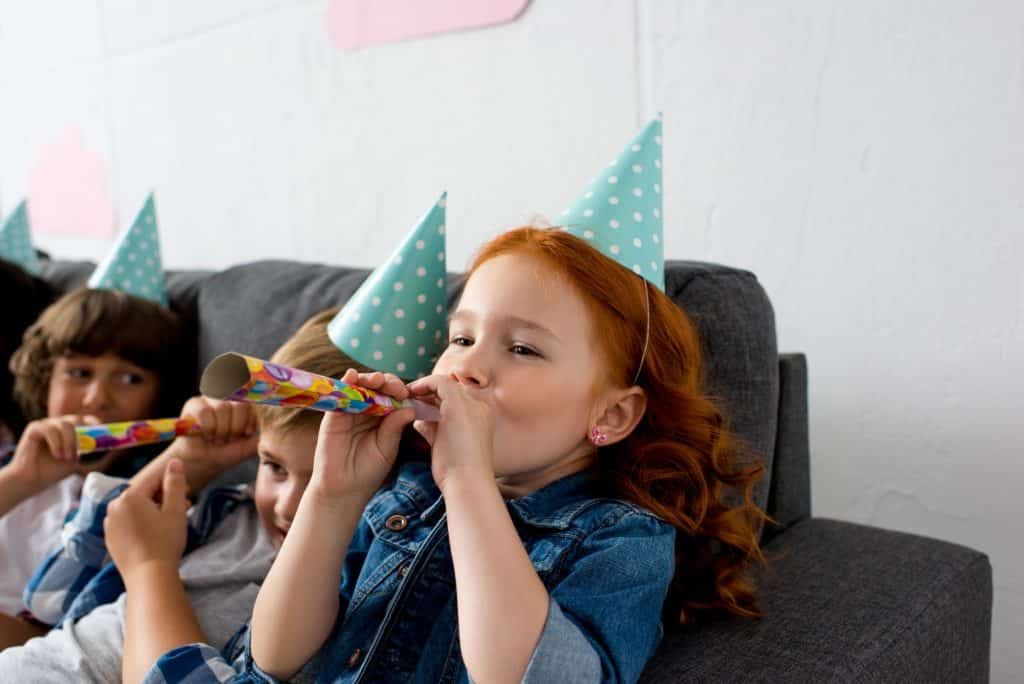 adorable little kids blowing in party blowers while sitting on sofa at birthday party