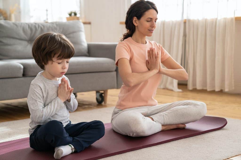 Mom with little son doing yoga exercise, practicing, teaching child to meditate, sit on mat at home.