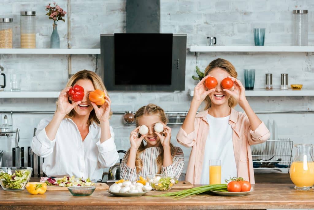 playful little child covering eyes with food with mother and grandmother and looking at camera while