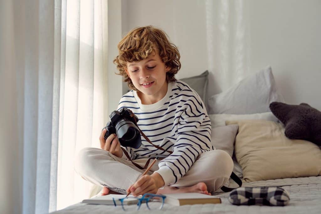 Schoolboy with photo camera writing in notepad on bed