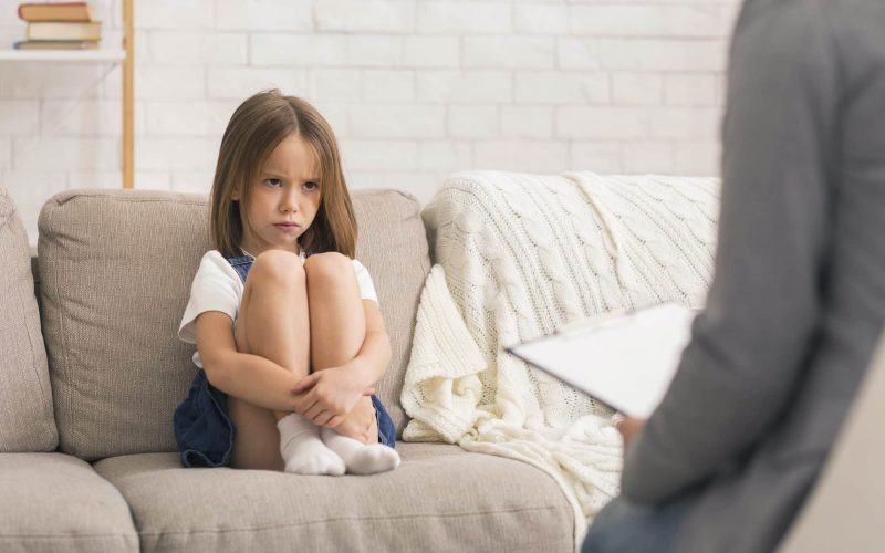 Concerned Little Girl Sitting Alone At Therapy Session With Children Psychologist