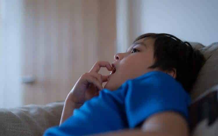 A boy sitting on sofa bitting finger nails while watching TV.Mixed race Child resting in living room