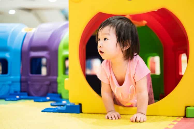 Little cute Asian toddler girl crawling out from colorful toy tunnel at playground.