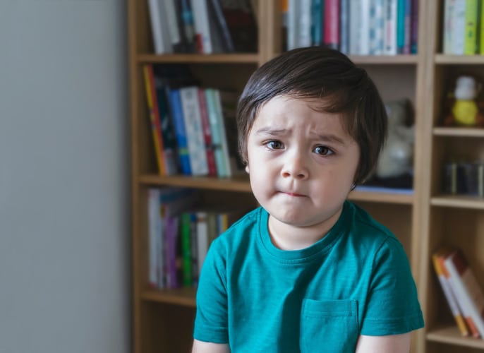 sad kid looking at camera with unhappy face, Angery little boy sitting alone with blurry bookshelf